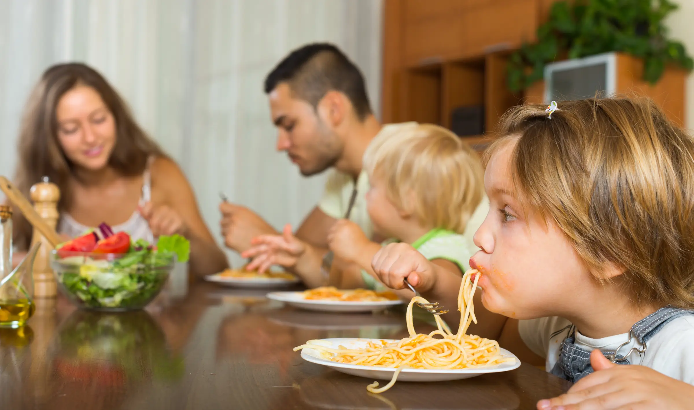 Familia almorzando espaguetis; un niño en primer plano sorbe la pasta